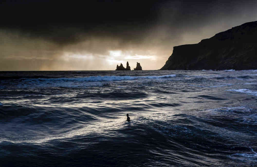 Small lone silhouetted figure stood low-chest deep in dark breaking sea near shore, with black skies overhead punctuated by horizon-brightness, and with black cliffs and pointed rock outcrops to the right.